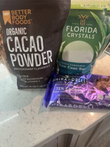 This image is of a white shiny counter top with grey flecks of color. On top of the counter is 3 packages; brown bag of organic cacao powder, a green bag of florida crystals powdered sugar, and a purple bag of 72% dark chocolate chips. 