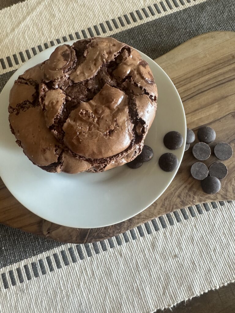 This image is of an off white table cloth, grey striped down the center and small grey lines on the edge. Wooden cutting board on top, topped with chocolate chips and a round, white, glass plate. On the plate is 2 chocolate cookies that are shiny and crinkled.