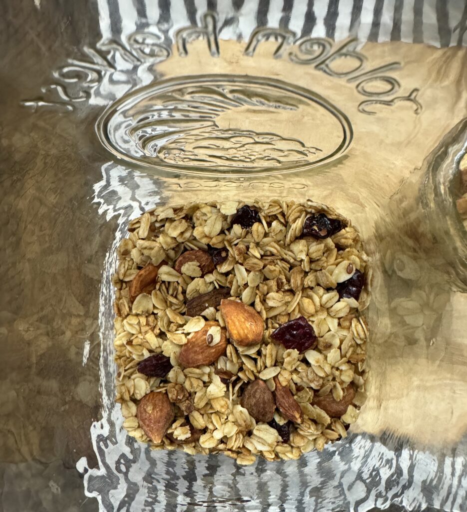 This is a picture of granola in the bottom of a glass jar. There are oats, red, dried cranberries, and almonds. The glass jar has an image at the top of the picture.