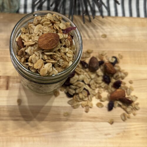 This picture is a wooden cutting board on top of a striped napkin. There is a glass jar sitting on top of the cutting board, full of granola, nuts and fruit. Some of the granola is sprinkled on the cutting board.