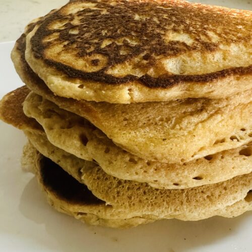 This image is of a stack of pancakes, light, golden brown. They are about 6 pancakes tall, stacked on a white plate.