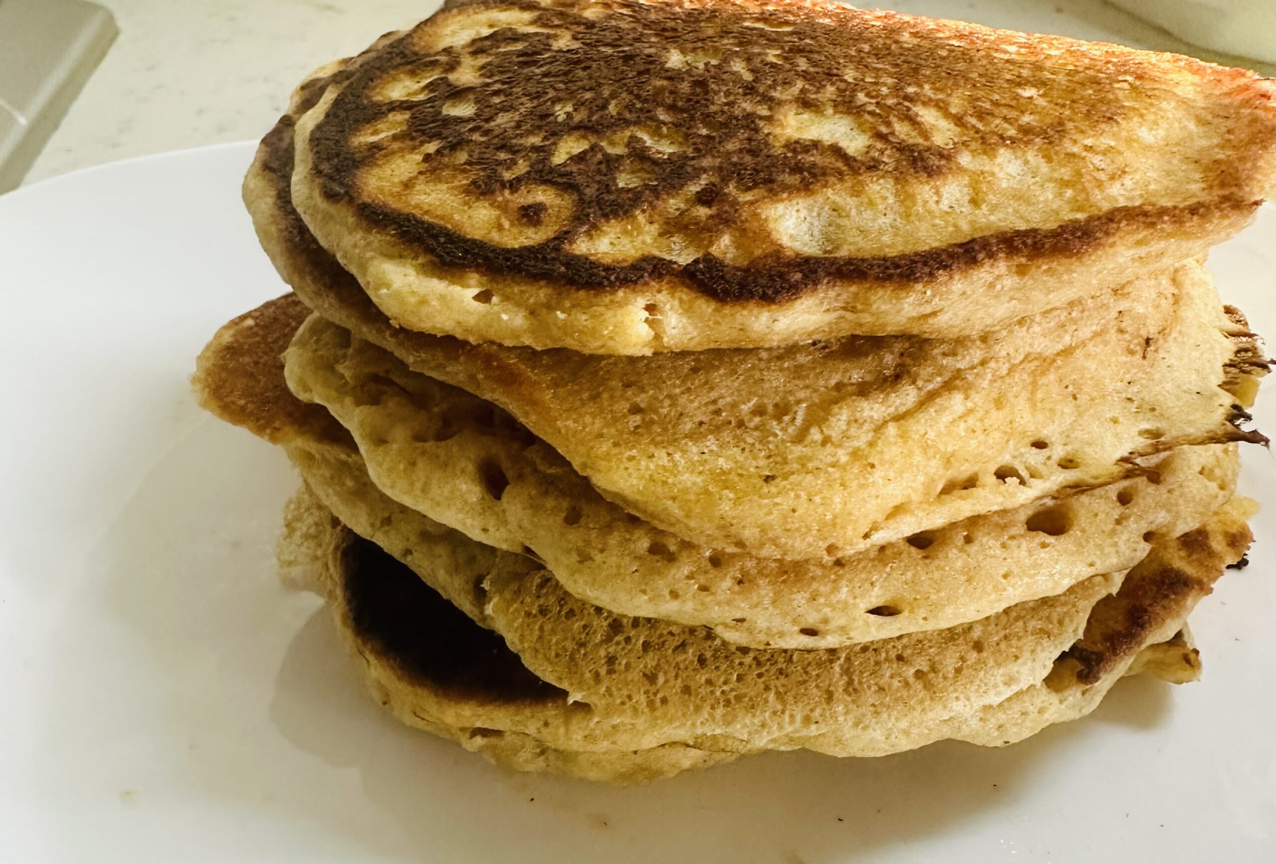 This image is of a stack of pancakes, light, golden brown. They are about 6 pancakes tall, stacked on a white plate.