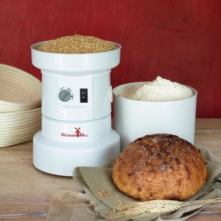 This is a picture of a white plastic grain mill in front of a red backdrop. It is full of wheat berries, and there is milled flour in the bowl next to it. There is a loaf of bread in front of the mill and white baskets behind the mill.