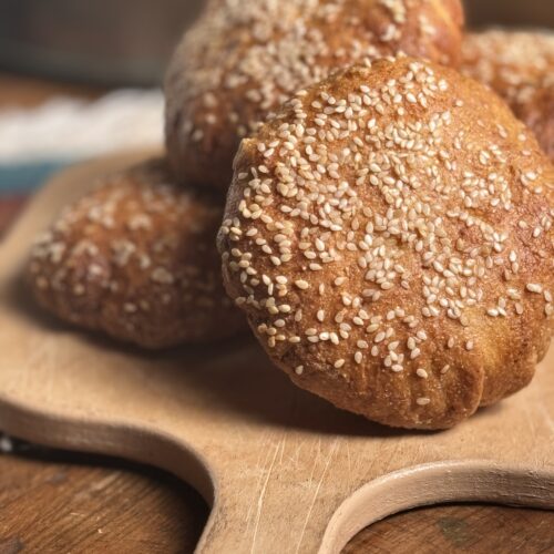 This image is of a light wooden cutting board on top of a dark wooden table. There is a stack of 5 golden brown hamburger buns topped with sesame seeds on top of it. There is a blue and white fringed pot holder in the background.