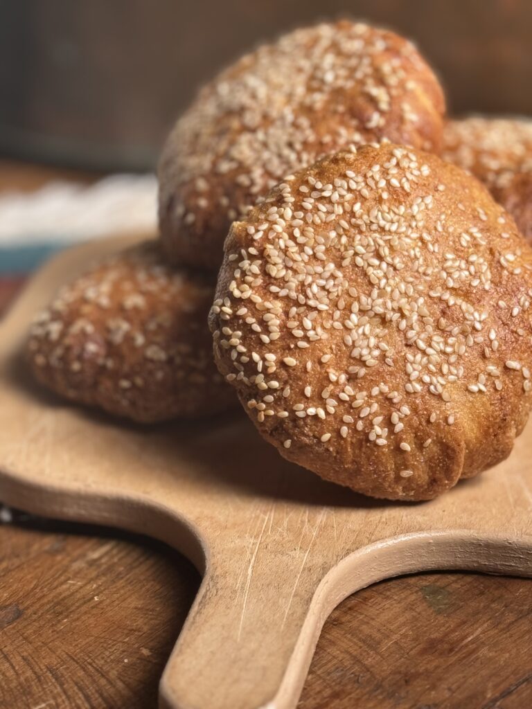 This image is of a light wooden cutting board on top of a dark wooden table. There is a stack of 5 golden brown hamburger buns topped with sesame seeds on top of it. There is a blue and white fringed pot holder in the background.