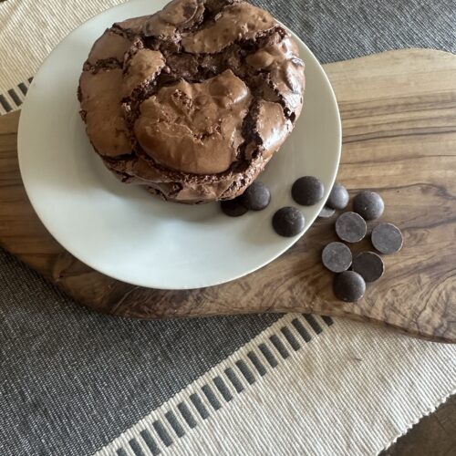 This image is of a wooden table, topped with an ivory table cloth with grey stipes and lines. The table cloth is topped with a wooden cutting board, sprinkled with chocolate chips and a white, glass plate. On the plate is a stack of 2 dark chocolate crinkle cookies, shiny brown with deep brown cracks.