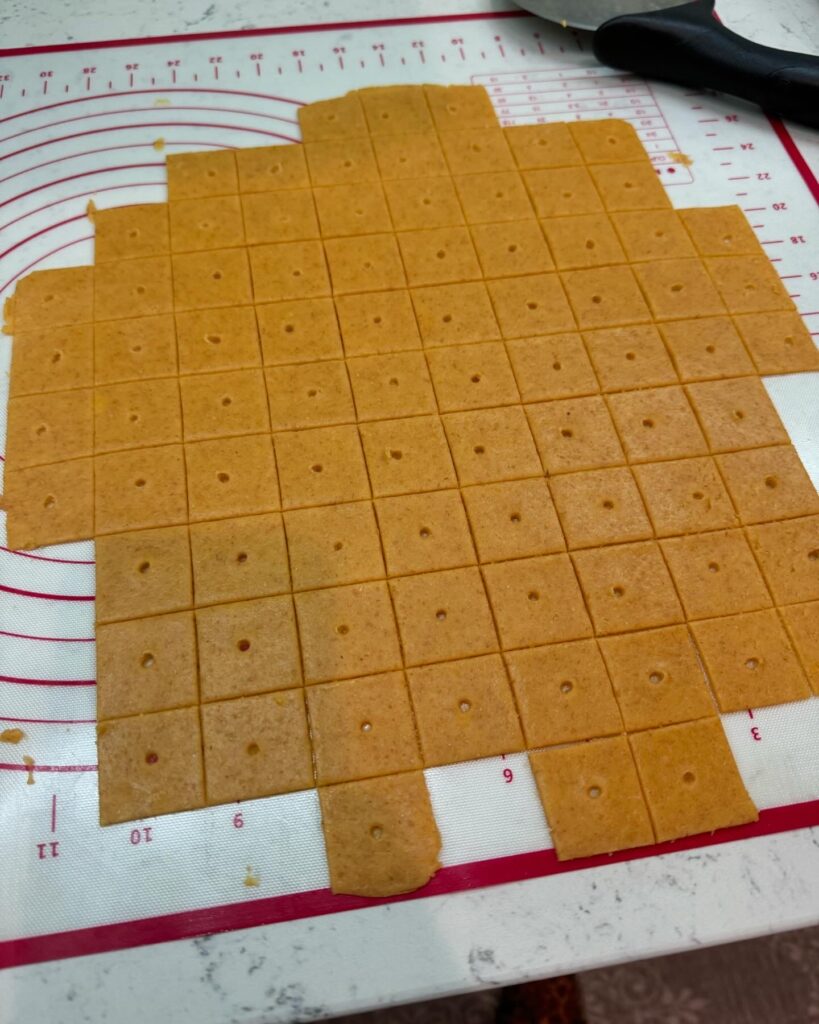 This image is of a white counter top, topped with a white silicone mat with red trim and markings. On the silicone mat is cheese crackers made with fresh milled flour. The crackers are rolled out thin, cut into small squares ad have a small hole in the center. There is a pizza cutter in the top of the image.