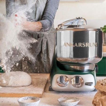 This image is of a green and stainless steel Ankarsrum Mixer next to a person in a blue shirt and grey apron, clapping flour between their hands causing it to puff out. There is a loaf of bread being formed on a wooden cutting board. This can also be found if you Shop Bailey Basic's Kitchen 