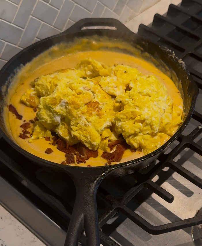 This image is of a cast iron skillet on the stove, filled with bacon egg and cheese mixture and a white tile background.