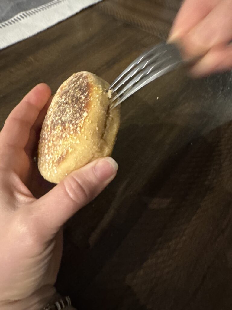 This image is of a hand holding and English Mufffin made with fresh milled flour being opened by inserting a fork and removing it. Moving around the outside of the muffin.