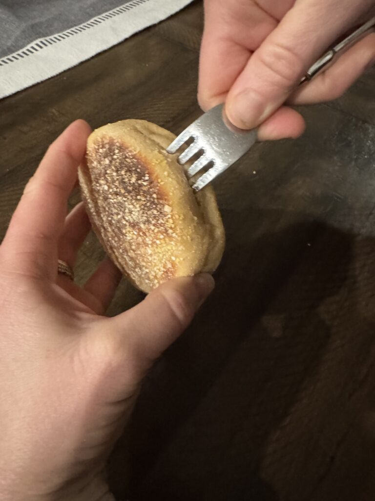 This image is of a hand holding and English Mufffin made with fresh milled flour being opened by inserting a fork and removing it. Moving around the outside of the muffin.