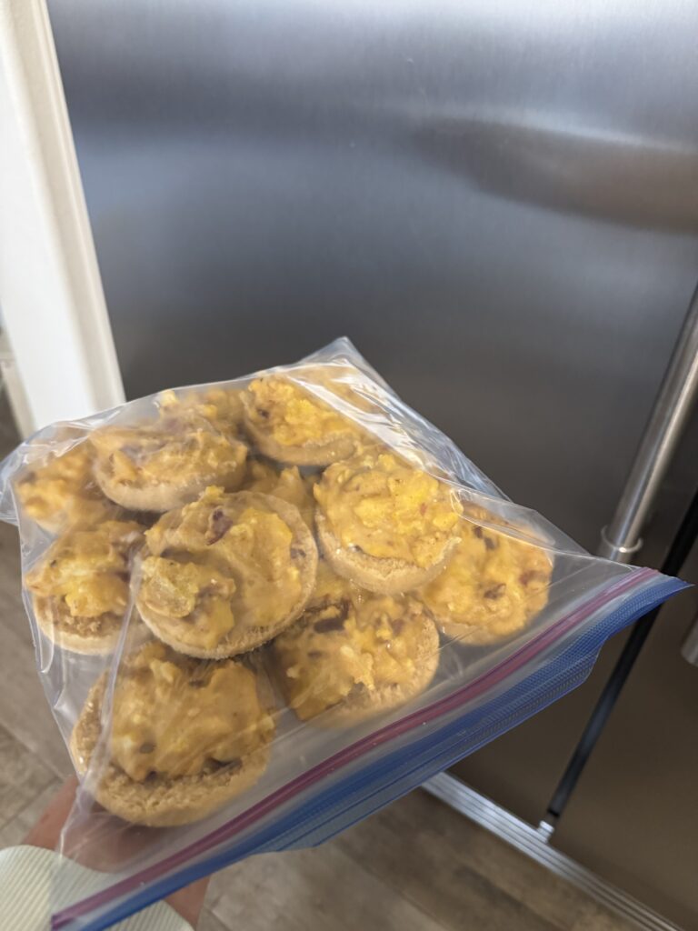 This image is of a gallon ziplock bag full of open faced english muffin breakfast sandwiches. The bag is in front of a large stainless steel freezer door.