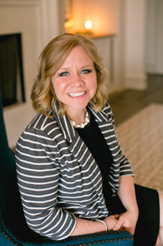 This image is of a lady with blonde hair, a lver necklace, a grey striped blazer and a black shirt. There is a tan rug in the background.