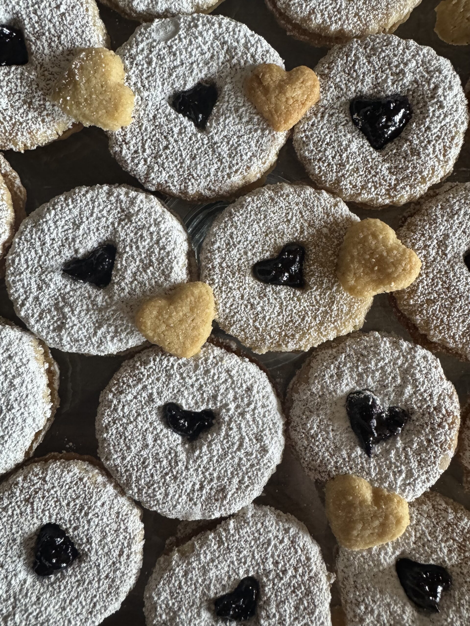 This image is of round cookies on a wooden table top. They have a heart cutout in the center and are dusted with powdered sugar. There are golden brown heart shaped cookies on top of the white cookies. There is dark jam coming through the heart cut outs.