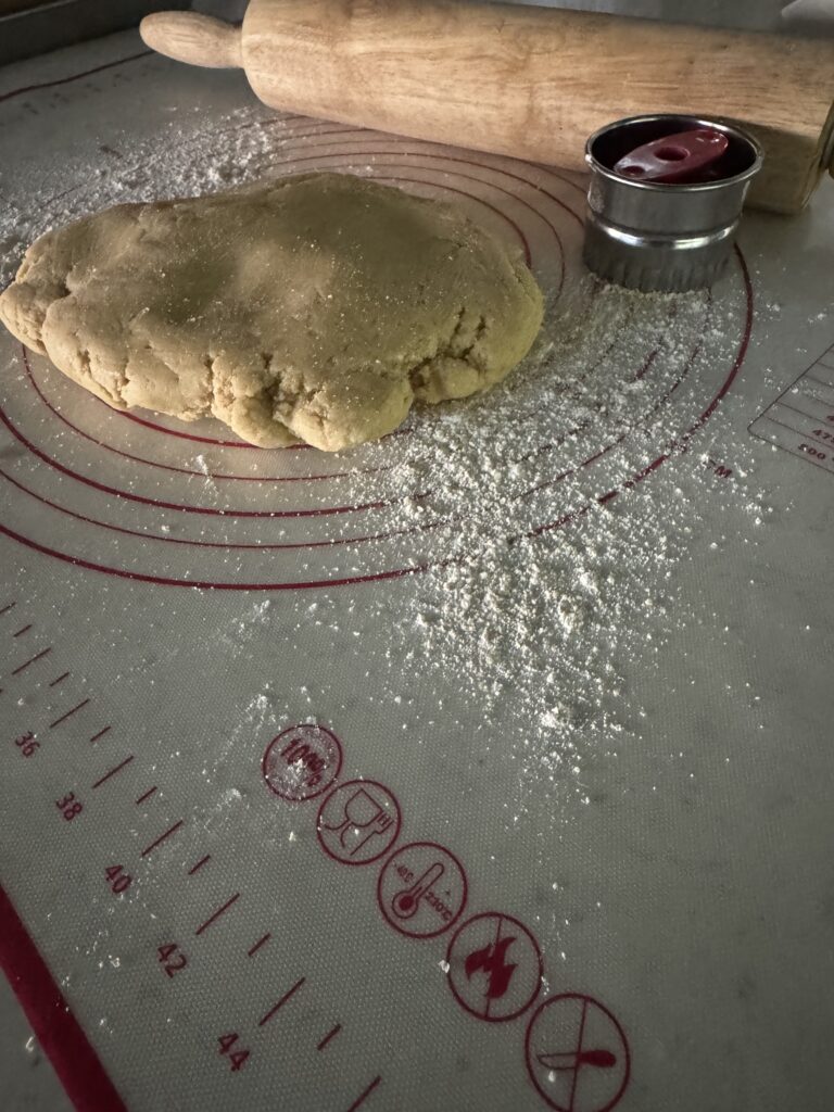 This image is of a white silicone mat with red circles and lines on it. There is white flour sprinkled on it, cookie dough in a ball, Linzer cookie cutter and rolling pin.