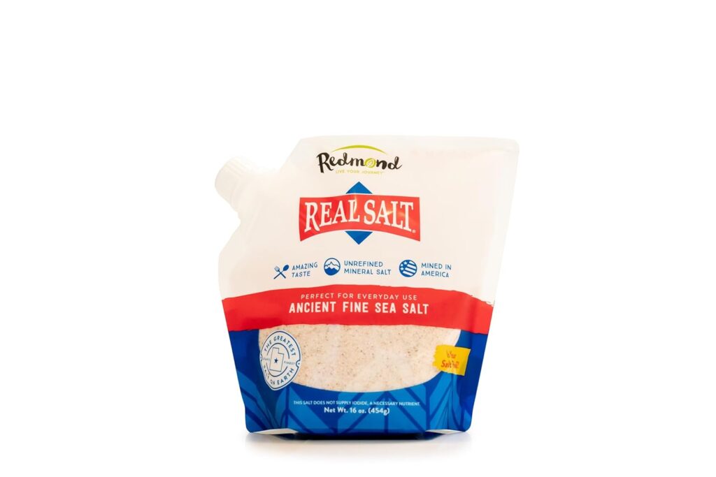 This image is of a bag of Redmond's real salt. The packagin is white at the top with a red line across the middle and a blue section at the bottom with a clear window to see the pink speckled salt. Bailey Basic's Kitchen NEVER runs out of this.