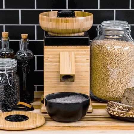 This image is of the NutriMill harvest mill. (vs. WonderMill) There is a black and white tile background, jars of grains and beans and a bamboo flour mill in the center. There is a black bowl in front to catch the flour.